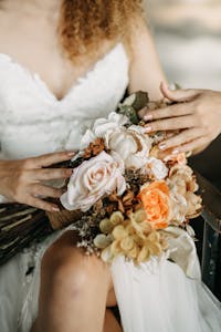 Close-up of a bride's delicate hands holding a floral bouquet in a wedding dress.