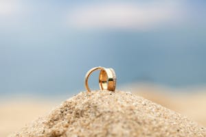 Close-up of elegant wedding rings placed on sand with ocean backdrop, evoking a romantic beach wedding setting.